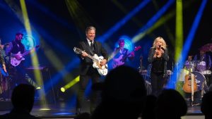 performer on stage with guitar, a singer, and a band with audience watching